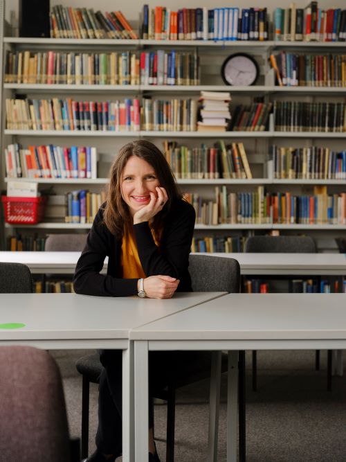 Foto von Dr. Yvonne Späck-Leigsnering, die vor einem Bücherregal an einem Schreibtisch sitzt und ihren Kopf mit der linken Hand abstützt