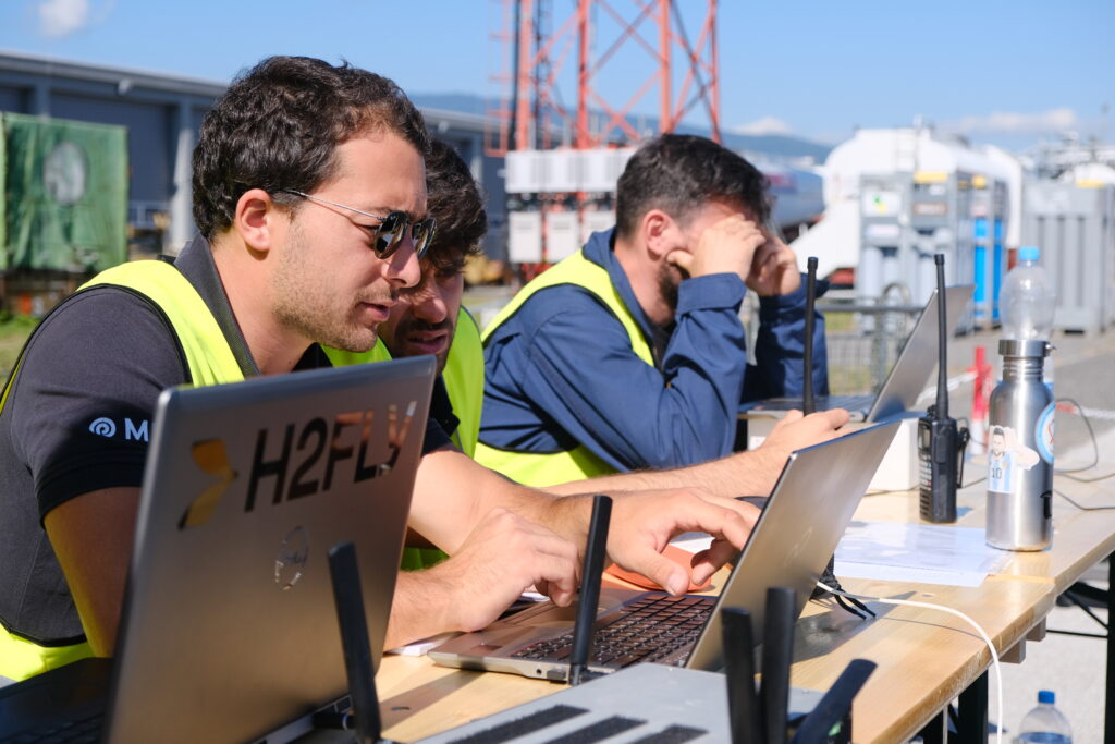 Drei Männer in Warnwesten sitzen auf einem Flugplatz vor drei Laptops