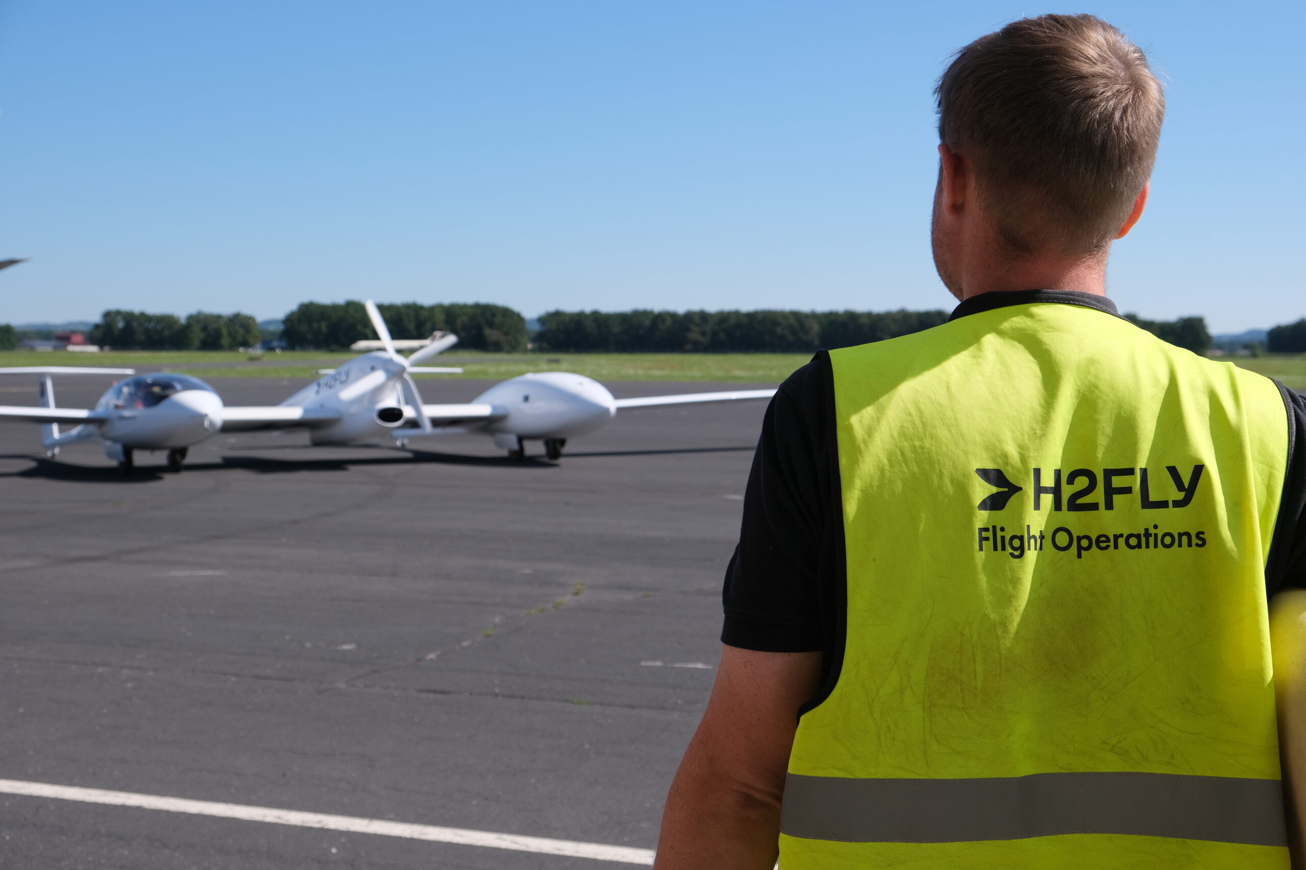 Foto eines Flugzeugs auf einem Flugplatz, links im Bild ist ein Mitarbeiter von hinten zu sehen, auf dessen Warnweste H2FLY steht