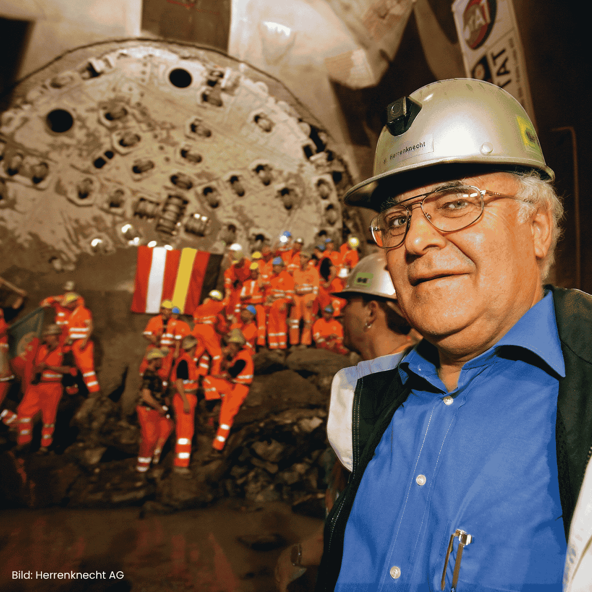 Foto von Martin Herreknecht mit Bauhelm vor einer Gruppe Arbeitender und einer Tunnelvortriebsmaschine am Gotthard-Basistunnel. Bild: Herrenknecht AG